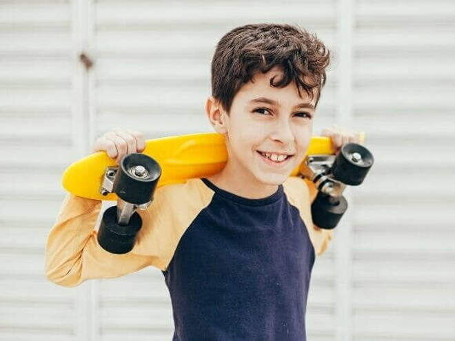 Menino sorrindo e segurando um skate pequeno em cima dos ombros