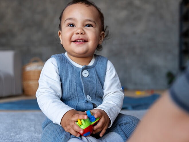 Bebê de aproximadamente 1 ano sentado no tapete da sala segurando brinquedos.