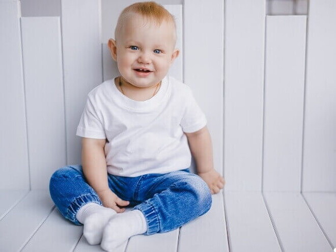 Um bebê sentado, sorrindo, veste uma camisa branca e uma calça jeans, sob um fundo branco.