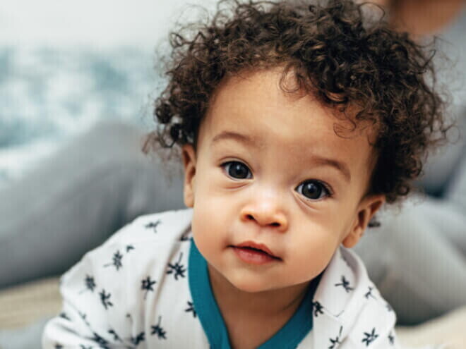 Bebê com cabelo cacheado vestindo um pijama branco com detalhes azuis, olhando diretamente para a câmera.