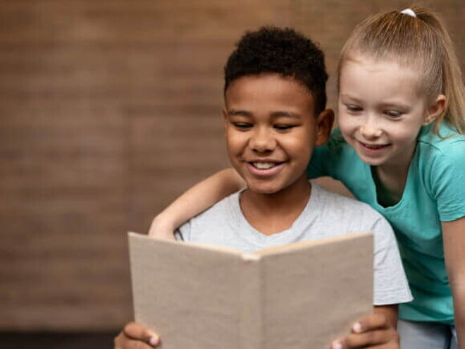 Duas crianças sorrindo, lendo um livro juntas.