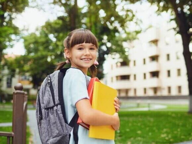 Uma menina sorrindo, segurando o material escolar e vestindo uniforme