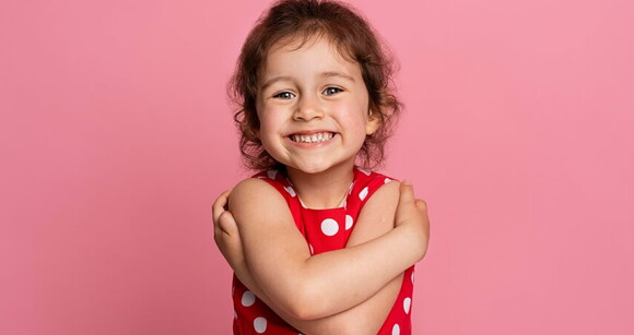 Menina usando roupa vermelha com bolinhas brancas sorrindo em frente a um fundo cor de rosa