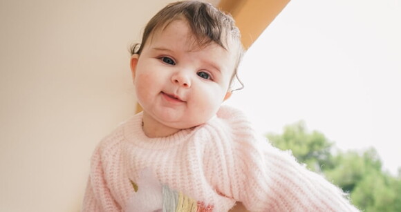 Bebê de olhos claros usando roupa de frio no colo da mãe.