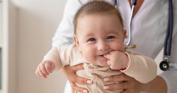 Bebê sorrindo com dedinhos na boca no colo de uma médica