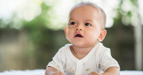 Bebê com macacão branco olhando atentamente com a boca aberta