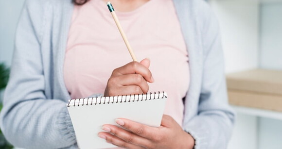 Mulher vestindo blusa rosa e casaco azul com caderno e lápis em mãos