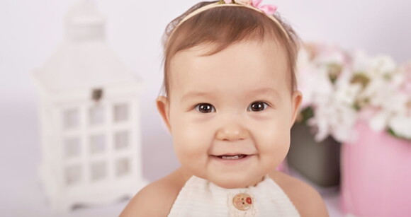 Menina de aproximadamente 1 ano usando vestido e tiara sorrindo.