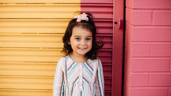 Menina sorrindo com um laço na cabeça em frente à uma parede amarela e vermelha.