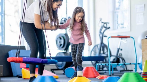 Uma mulher sorrindo e segurando a mão de uma criança, que está andando em um espaço para exercícios infantis.