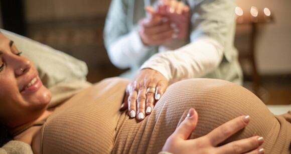 Mulher grávida deitada em uma cama recebendo o toque suave de uma doula em sua barriga