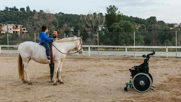 Criança montada em um cavalo ao lado de um homem, com uma cadeira de rodas à frent