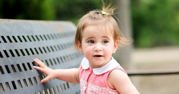 Menina de aproximadamente 2 anos usando vestido cor de rosa sentada no banco de um parque.