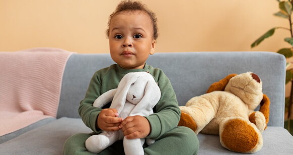 Bebê de cabelo cacheado sentado no sofá usando pijama e abraçando seus ursinhos de pelúcia.