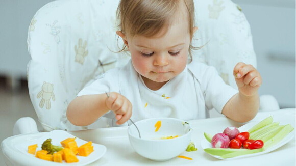 Um bebê sentado em uma cadeira alta para alimentação, brincando com uma colher e comendo pedaços de vegetais.