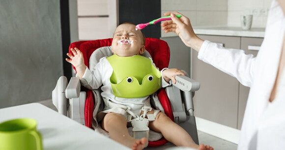 Mãe tentando dar papinha para bebê que está na cadeirinha chorando.