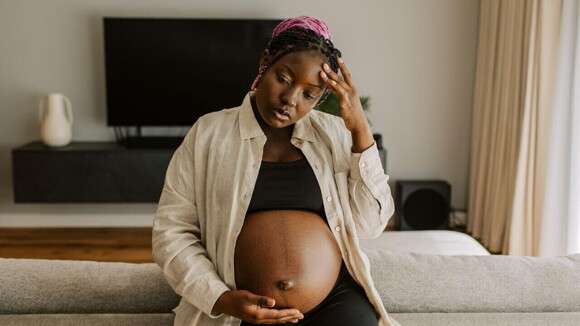 Mulher grávida com uma camisa bege, sentada com uma mão sobre a barriga e outra na testa, aparentando estar desconfortável.