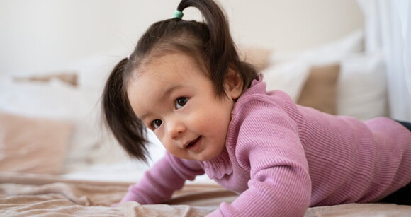 Bebê asiática usando roupa cor de rosa engatinhando em cima da cama.