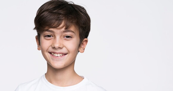 Menino de cabelo liso usando camiseta branca e sorrindo em frente a uma parede branca.