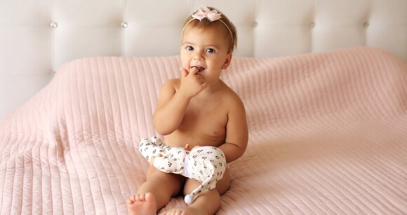 Menina de aproximadamente 1 ano sentada na cama brincando com uma boneca.