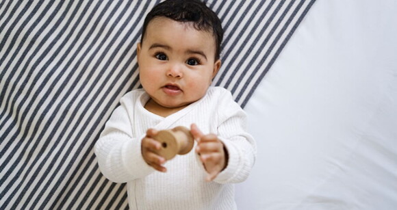 Bebê deitado em uma cama vestindo roupa branca e segurando um brinquedo de madeira