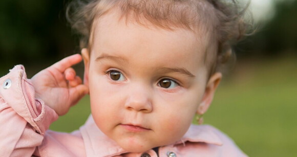 Bebê usando roupa cor de rosa e passando a mão na orelha.