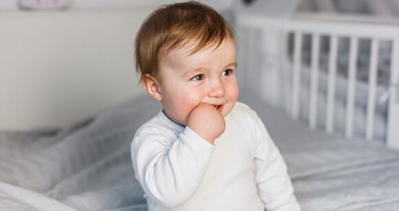 Menino pequeno de aproximadamente 1 ano sentado na cama com a mão na boca.