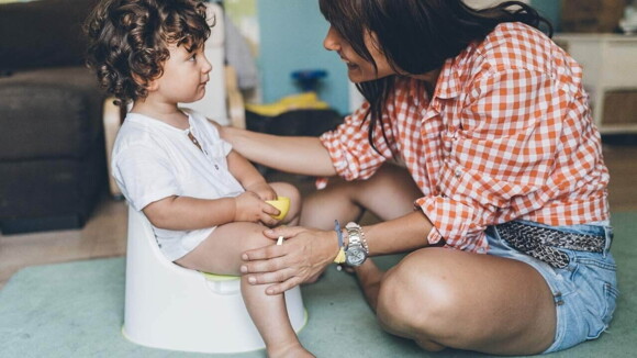 Bebê de cabelos cacheados sentado em um penico branco, e sua mãe sentada em sua frente usando camisa xadrez e short jeans.