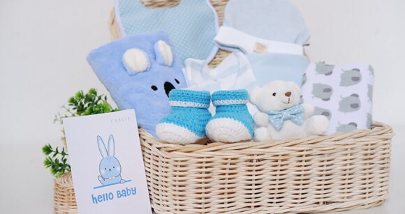 Imagem de uma cesta com vários itens de bebê, como cobertor, manta, sapatinho, babador, touca e um coelhinho de pelúcia 