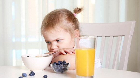 Criança sentada em uma mesa com a cabeça deitada sobre os braços, com olhar irritado, observando um pote de comida à sua frente.