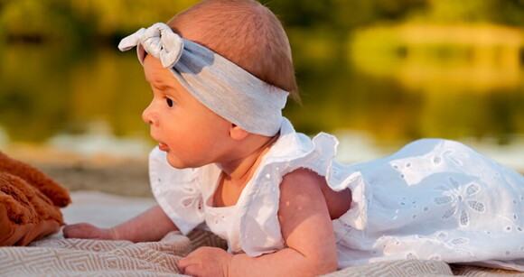 Bebê deitada de bruços usando faixa na cabeça e vestido branco