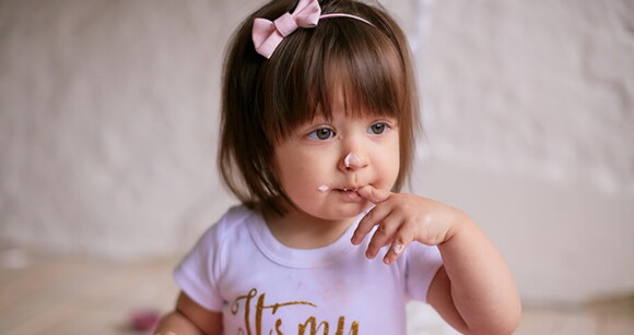 Menina de aproximadamente dois anos de olhos claros usando tiara de lacinho rosa e com o dedo na boca.