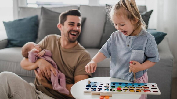 Um homem sorrindo e sentado no chão com um bebê no colo, ao lado de uma criança em pé e sorrindo, brincando com objetos sobre uma mesa.