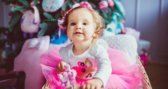 Menina de aproximadamente 1 ano usando vestido rosa e segurando um ursinho de pelúcia.