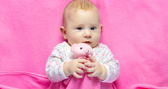 Bebê deitada na cama usando roupa cor de rosa e abraçando ursinho de pelúcia.