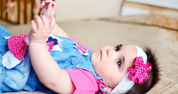 Bebê usando vestido jeans com flores cor de rosa e faixa na cabeça deitada de barriga para cima.