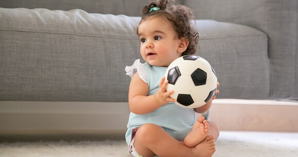 Bebê sentada em um tapete e segurando uma bola de futebol