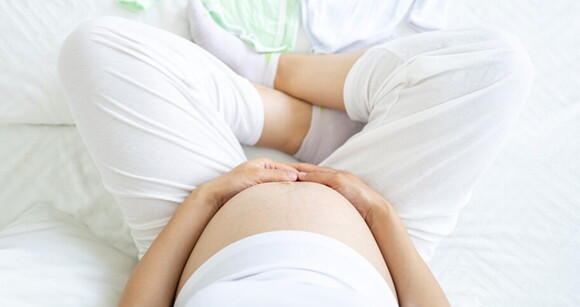 mulher com roupa branca sentada na cama enquanto segura a barriga de grávida