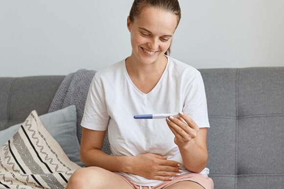 Mulher sentada no sofá com semblante de felicidade olhando para um teste de gravidez