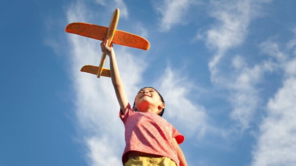 Com o céu de fundo, um menino brinca com um avião vestindo uma camisa rosa.