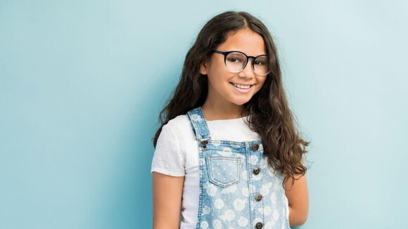 Menina de cabelos pretos e longos usando óculos e macacão jeans em frente a uma parede azul claro.