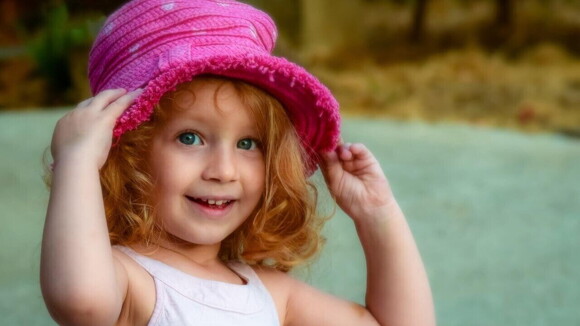 Menina sorrindo com as mãos em um chapéu rosa sobre a sua cabeça