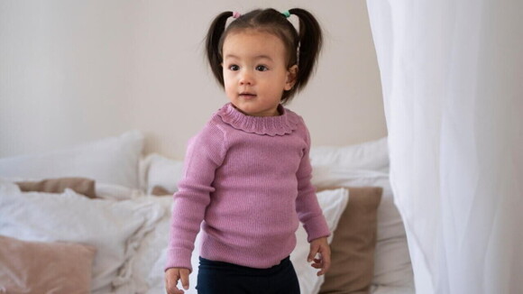 Uma menina de aproximadamente 2 anos, usando uma blusa lilás e calça preta, posando em um quarto.