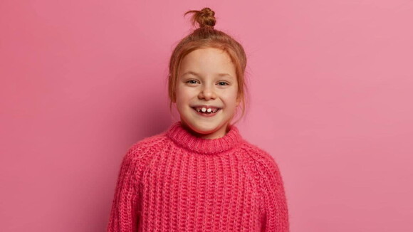 Uma menina de aproximadamente 6 anos sorrindo, com um suéter rosa, de frente para uma parede rosa.