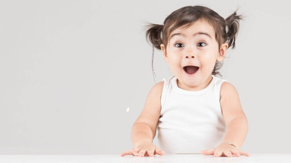 Menina sorridente com os olhos atentos usando uma blusa branca e com as mãos apoiadas sobre uma superfície.