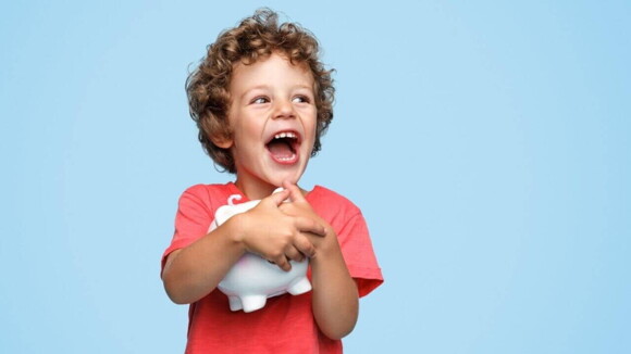 Menino sorridente com os cabelos cacheados abraçando um cofrinho branco em um fundo azul.