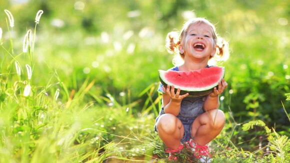 Menina sorridente agachada sobre a grama segurando uma melancia.