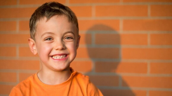 Menino vestindo uma camiseta laranja e sorrindo em frente a uma parede de tijolos