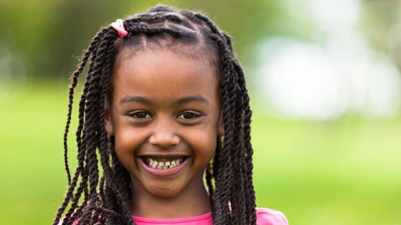 Menina com tranças e blusa rosa sorrindo e olhando diretamente para a câmera.