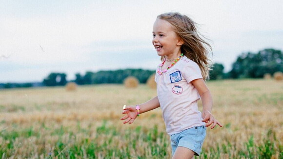 Menina sorridente de blusa cor de rosa e short jeans correndo em um campo verde.